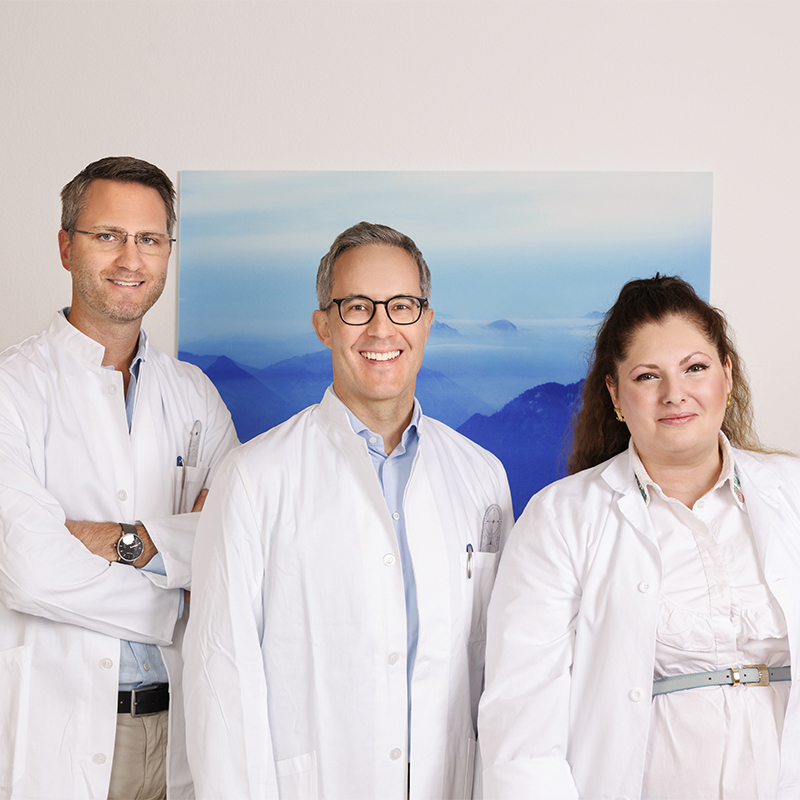 Foto des Ärzte-Teams von Orthopädie Wüst vor einem blauen Bild, mit Dr. med. Daniel Burger, Dr. med. Daniel Wüst und Dr. med. Patricia Scortia